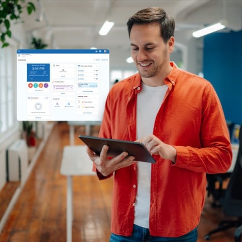 A person stands in a modern office holding a tablet, with a digital dashboard interface floating beside them and desks, chairs, and plants visible in the background