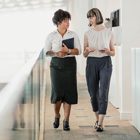 two women walking in a professional office setting carrying mobile phone and tablet